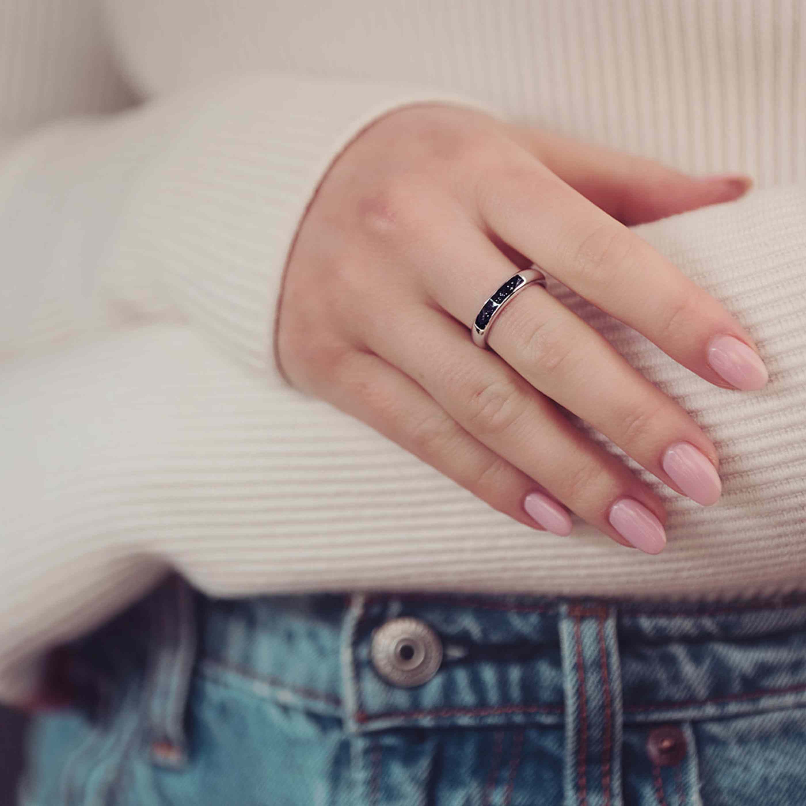 close up a womens hand Womens Cremation Ashes Ring Band, filled with ashes and black crushed crystal. The lady has pink painted nails and is wearing a white jumper