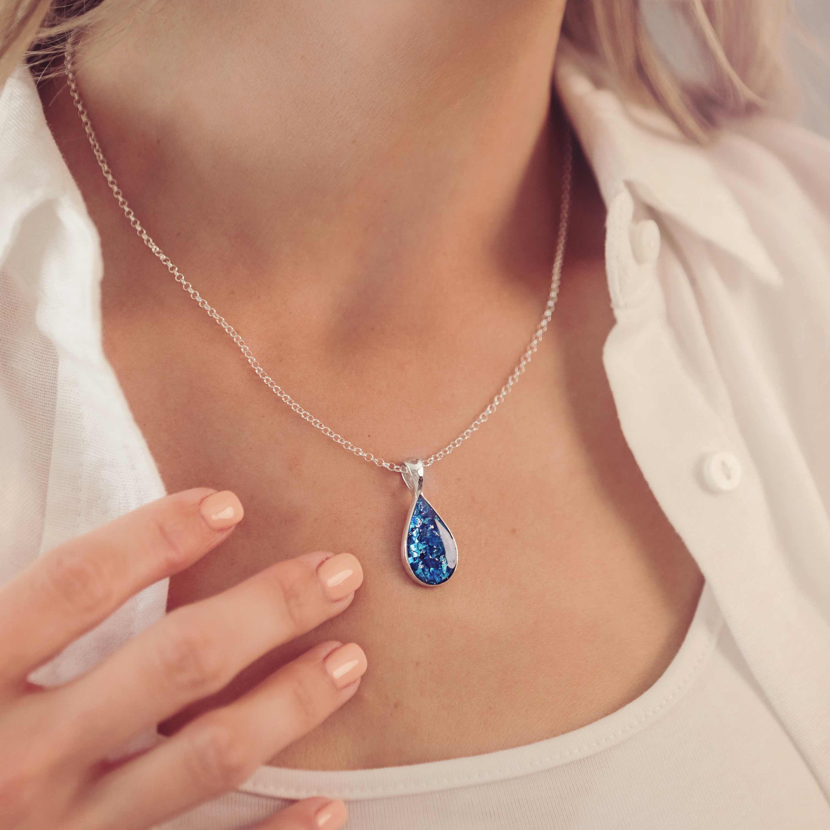 lady wearing a blue teardrop shaped ashes necklace filled with cremation ashes, she is wearing a white shirt and has beige painted nails