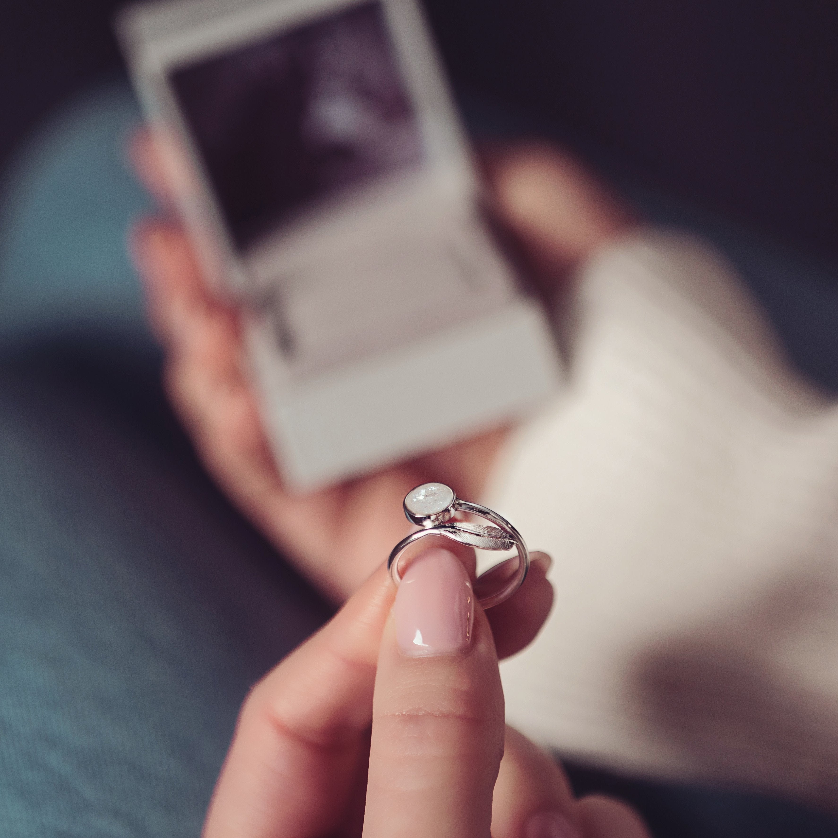 Hand holding a silver memorial feather ring containing cremation ashes. In the background, an open jewellery box is blurred