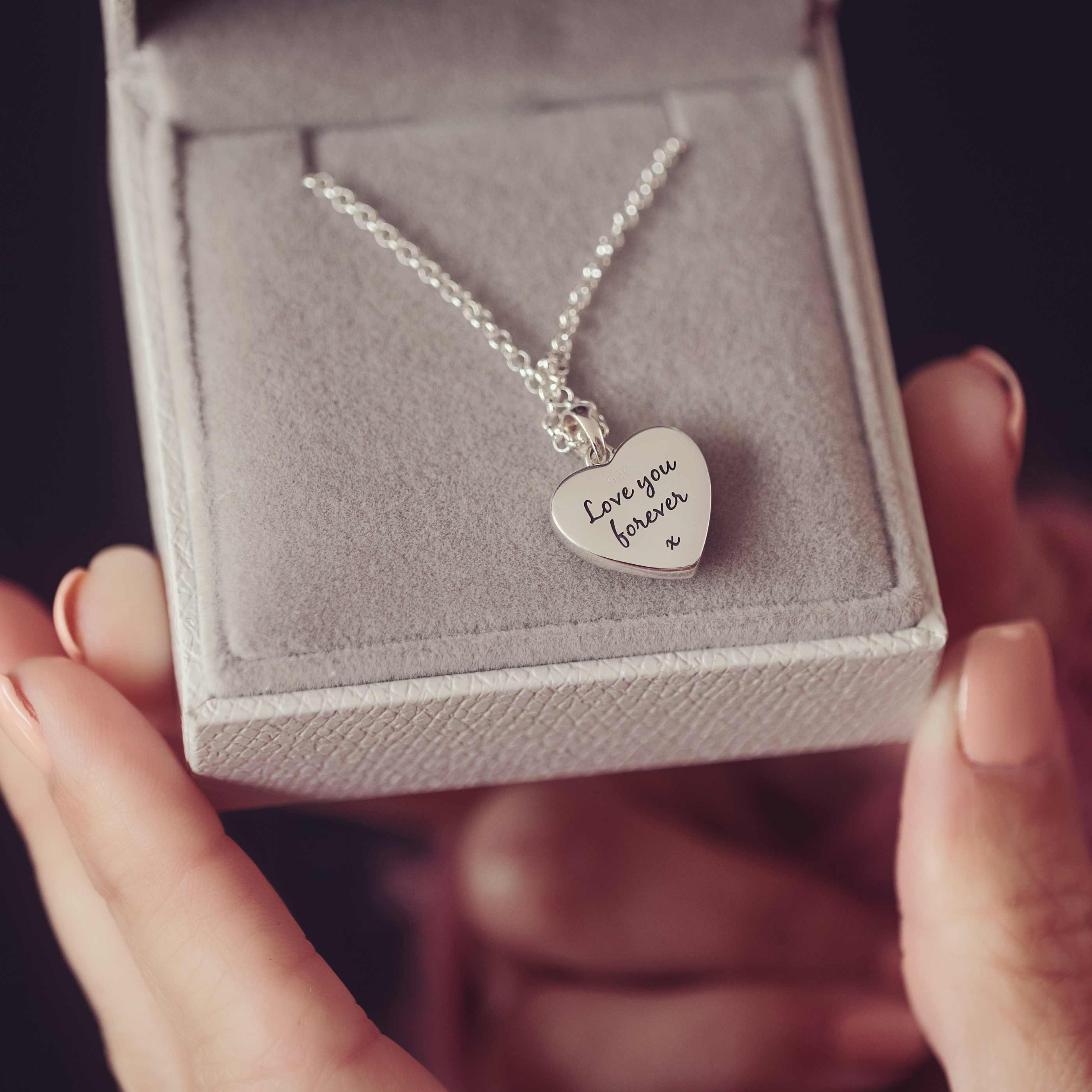 A close up shot of a lady's hands holding a grey jewellery box. Inside the box is a silver heart necklace with engraving. 