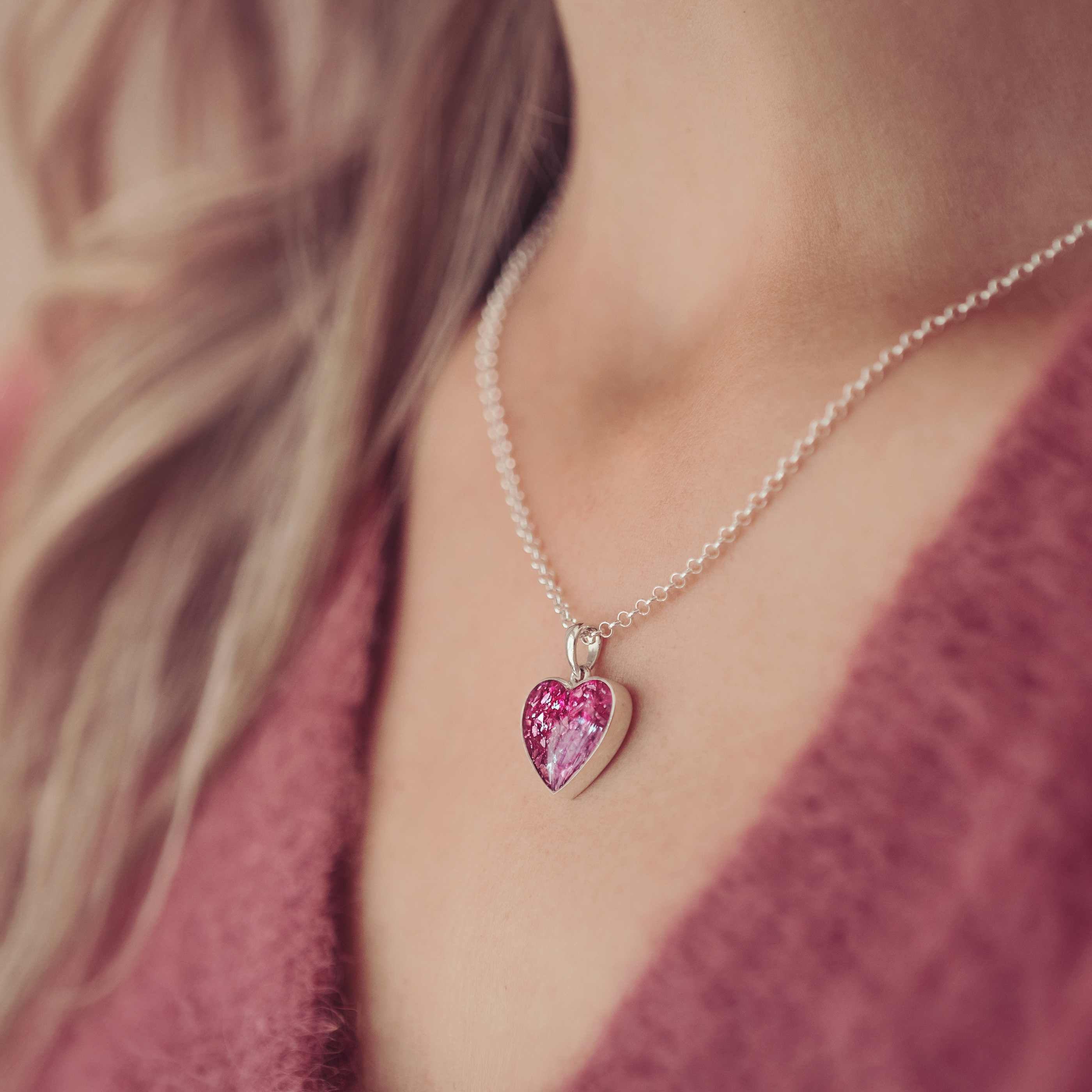 image cropped into a lady's neck showing a small silver and pink heart necklace. The necklace is filled with cremation ashes. 