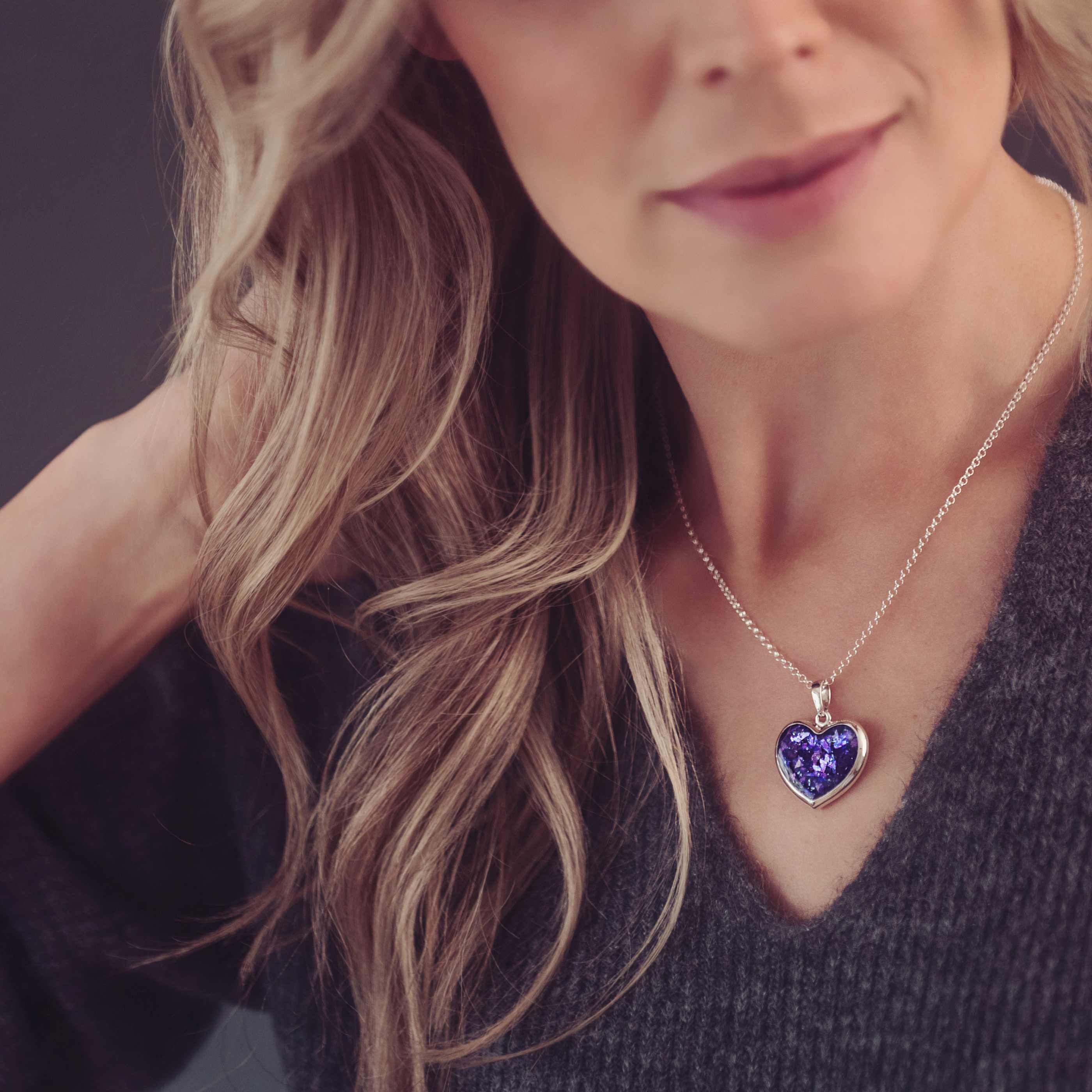 Lady with blonde hair wearing a grey knitted jumper and purple necklace filled with memorial cremation ashes