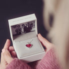 A red heart cremation ashes necklace being held in a grey jewellery box with a photograph of three people. 