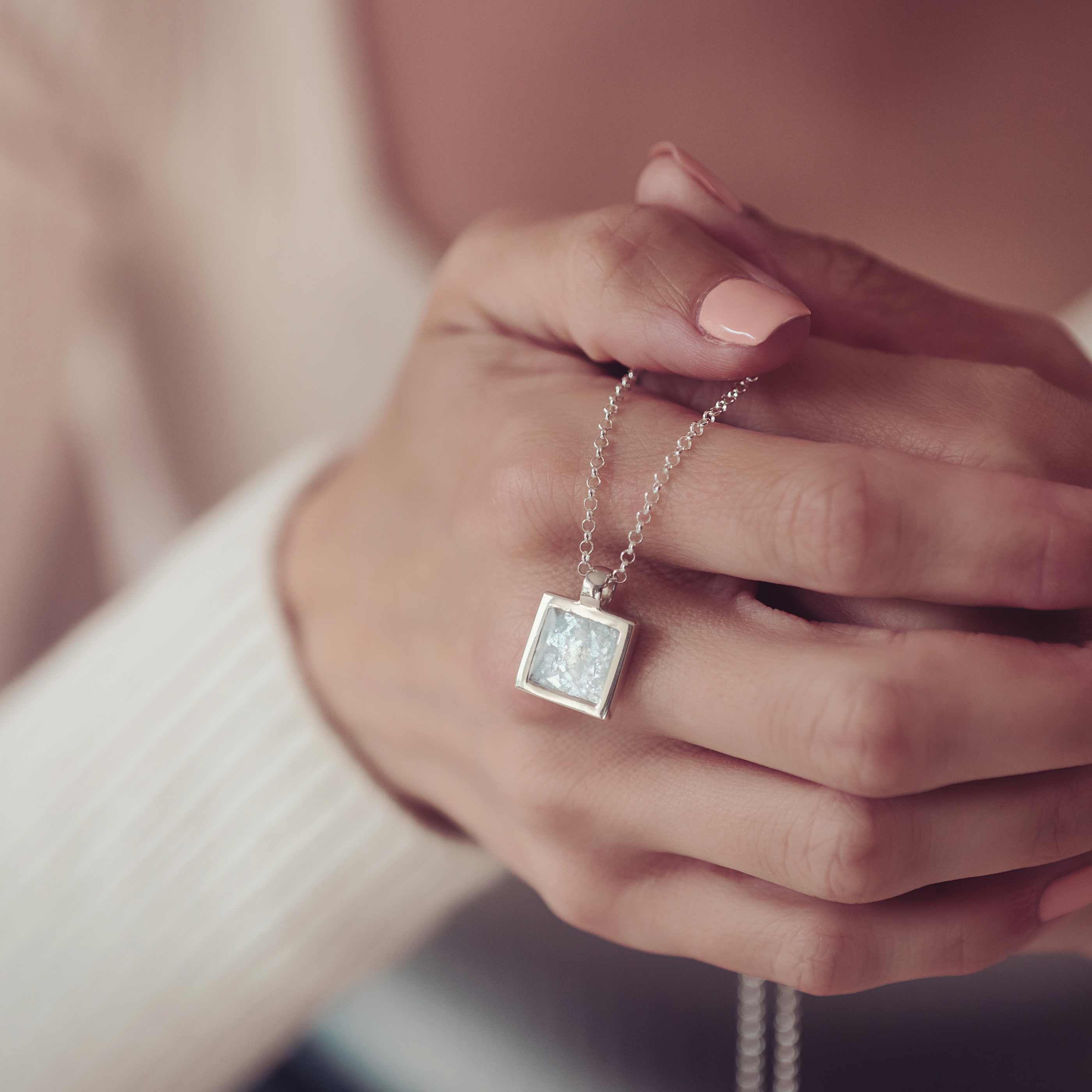 The image shows a close-up of a person's hands gently holding a necklace with a square-shaped silver pendant. The pendant contains a light-coloured, sparkling material, possibly ashes or resin, suspended on a delicate silver chain. The person's nails are neatly manicured in soft pink, and they are wearing a light-coloured ribbed sweater. The image focuses on the pendant, suggesting its sentimental or memorial significance, with soft tones and gentle lighting creating a calm and intimate atmosphere.