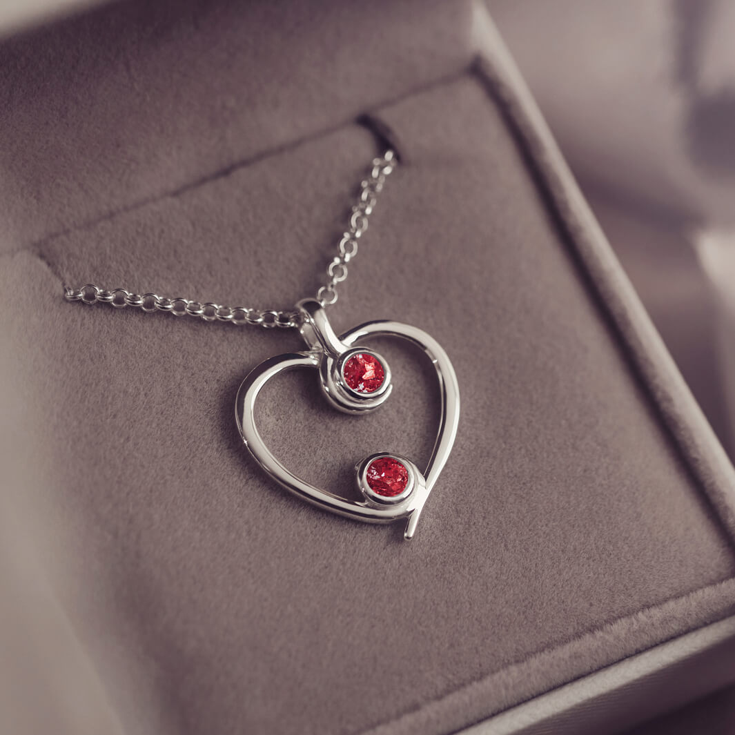 A silver heart-shaped pendant necklace featuring two sparkling red resin stones, holding cremation ashes, displayed against a neutral background.