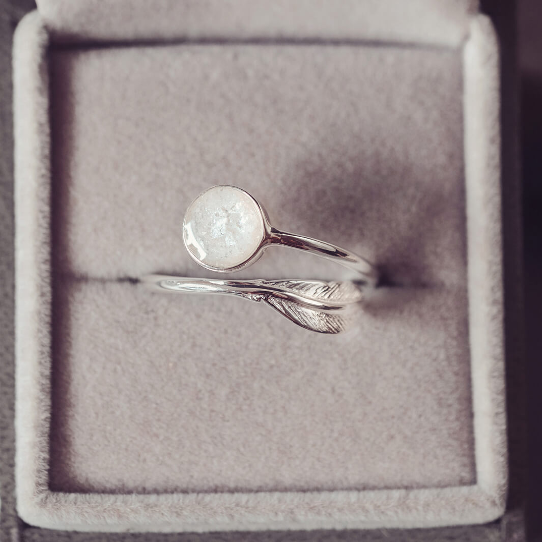 A sterling silver adjustable ring, with a circular bezel filled with clear and white crystals and ashes and a silver feather embellishment