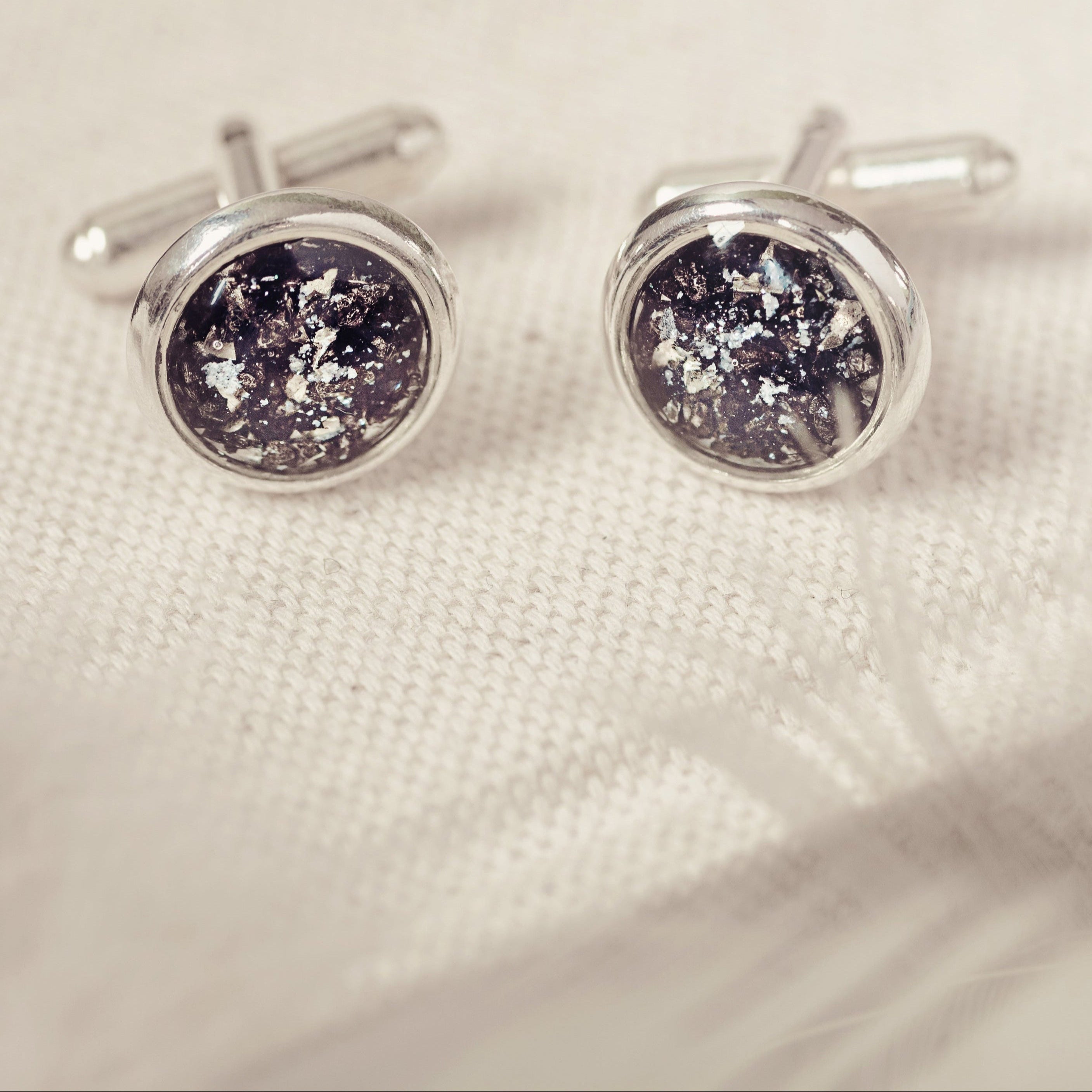 A pair of silver cremation ashes cufflinks with black shimmering resin, delicately placed on a soft, neutral fabric with wisps of feathers