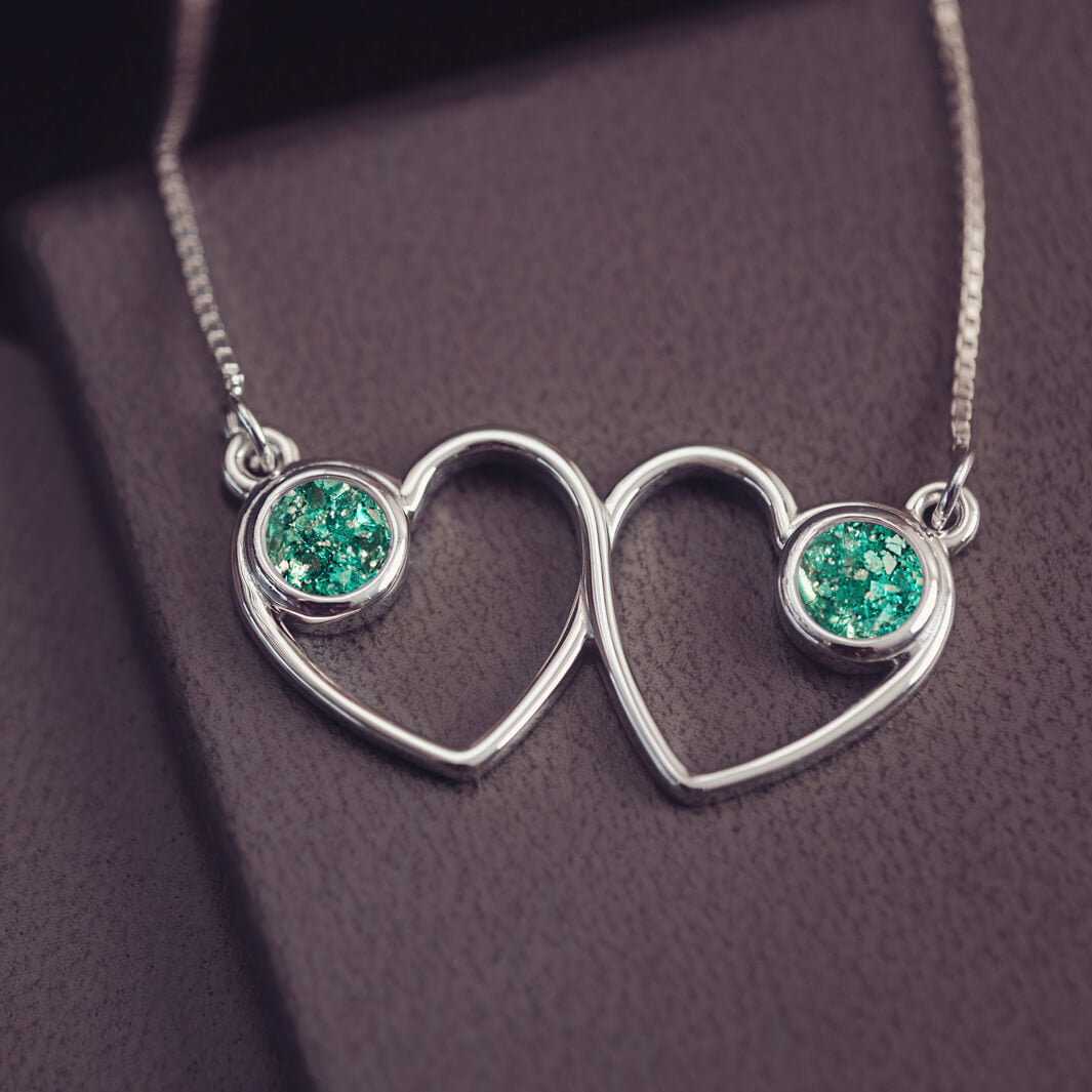 A delicate silver necklace featuring two intertwined heart-shaped pendants, each with a green resin stone which holds crystal and cremation ashes. Displayed against a neutral background.