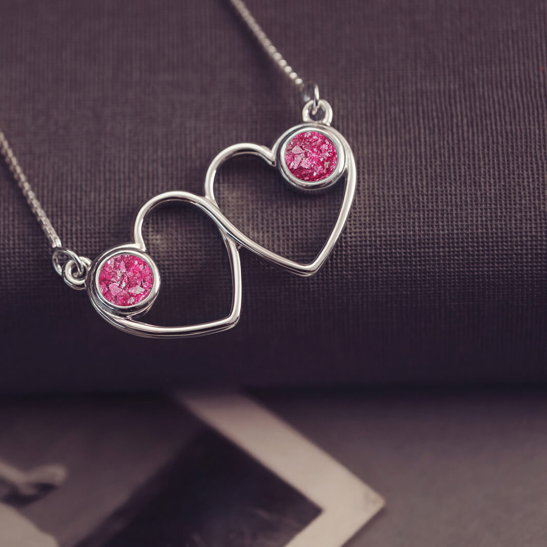 A delicate silver necklace featuring two intertwined heart-shaped pendants, each with a pink resin stone which holds crystal and cremation ashes. Displayed against a neutral background.