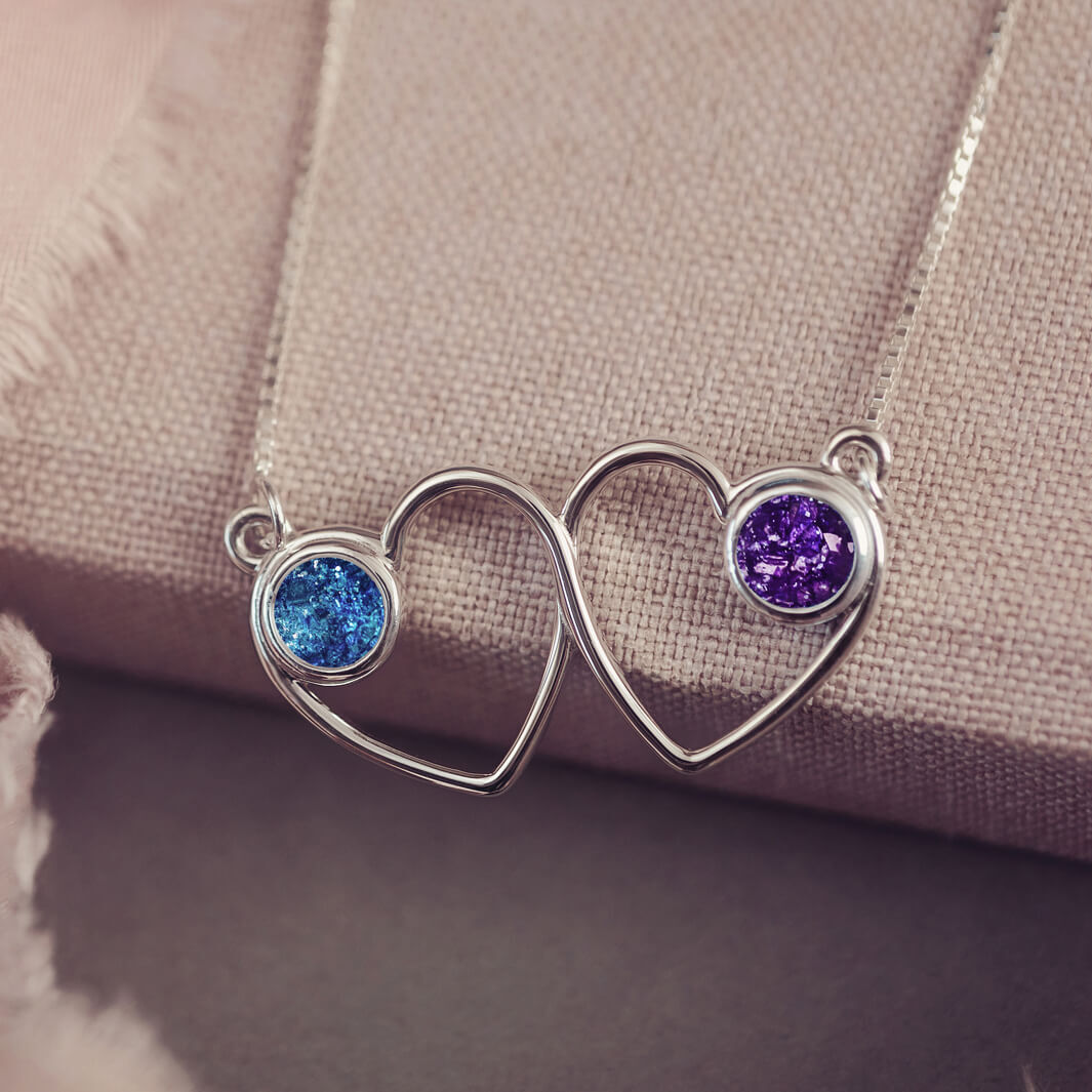 A delicate silver necklace featuring two intertwined heart-shaped pendants, each with a blue and purple resin stone which holds crystal and cremation ashes. Displayed against a neutral background.