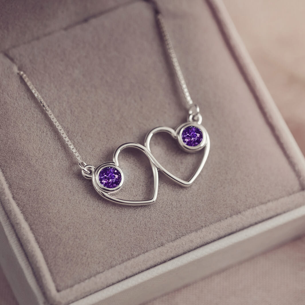 A delicate silver necklace featuring two intertwined heart-shaped pendants, each with a purple resin stone which holds crystal and cremation ashes. Displayed against a neutral background.