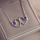 A delicate silver necklace featuring two intertwined heart-shaped pendants, each with a purple resin stone which holds crystal and cremation ashes. Displayed against a neutral background.