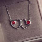 A delicate silver necklace featuring two intertwined heart-shaped pendants, each with a red resin stone which holds crystal and cremation ashes. Displayed against a neutral background.