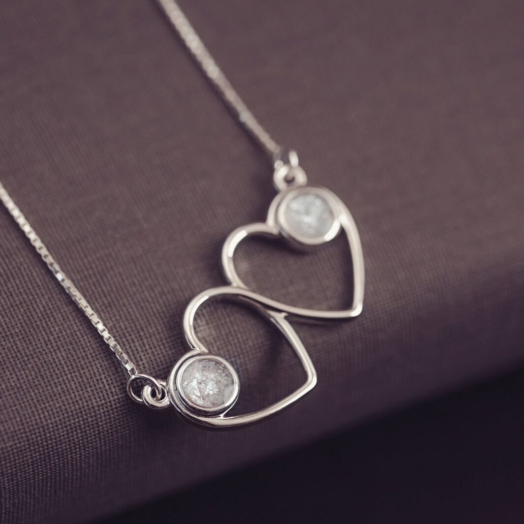 A delicate silver necklace featuring two intertwined heart-shaped pendants, each with a white resin stone which holds crystal and cremation ashes. Displayed against a neutral background.