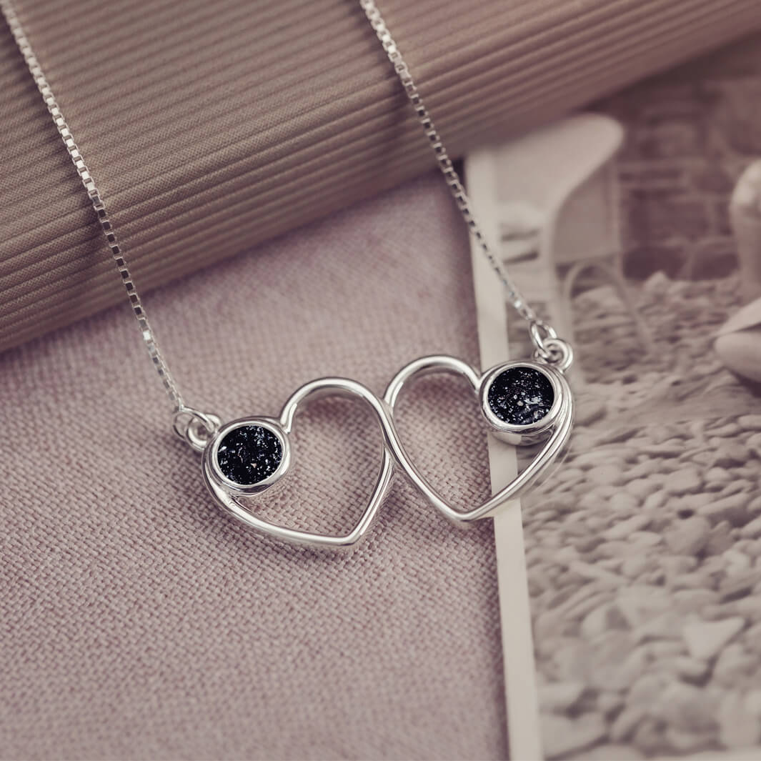 A delicate silver necklace featuring two intertwined heart-shaped pendants, each with a black resin stone which holds crystal and cremation ashes. Displayed against a neutral background.