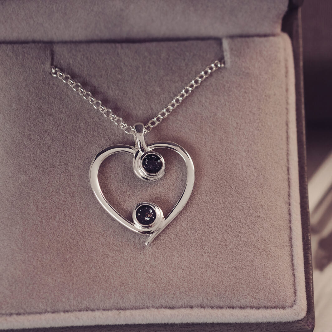 A silver heart-shaped pendant necklace featuring two sparkling black resin stones, holding cremation ashes, displayed against a neutral background.