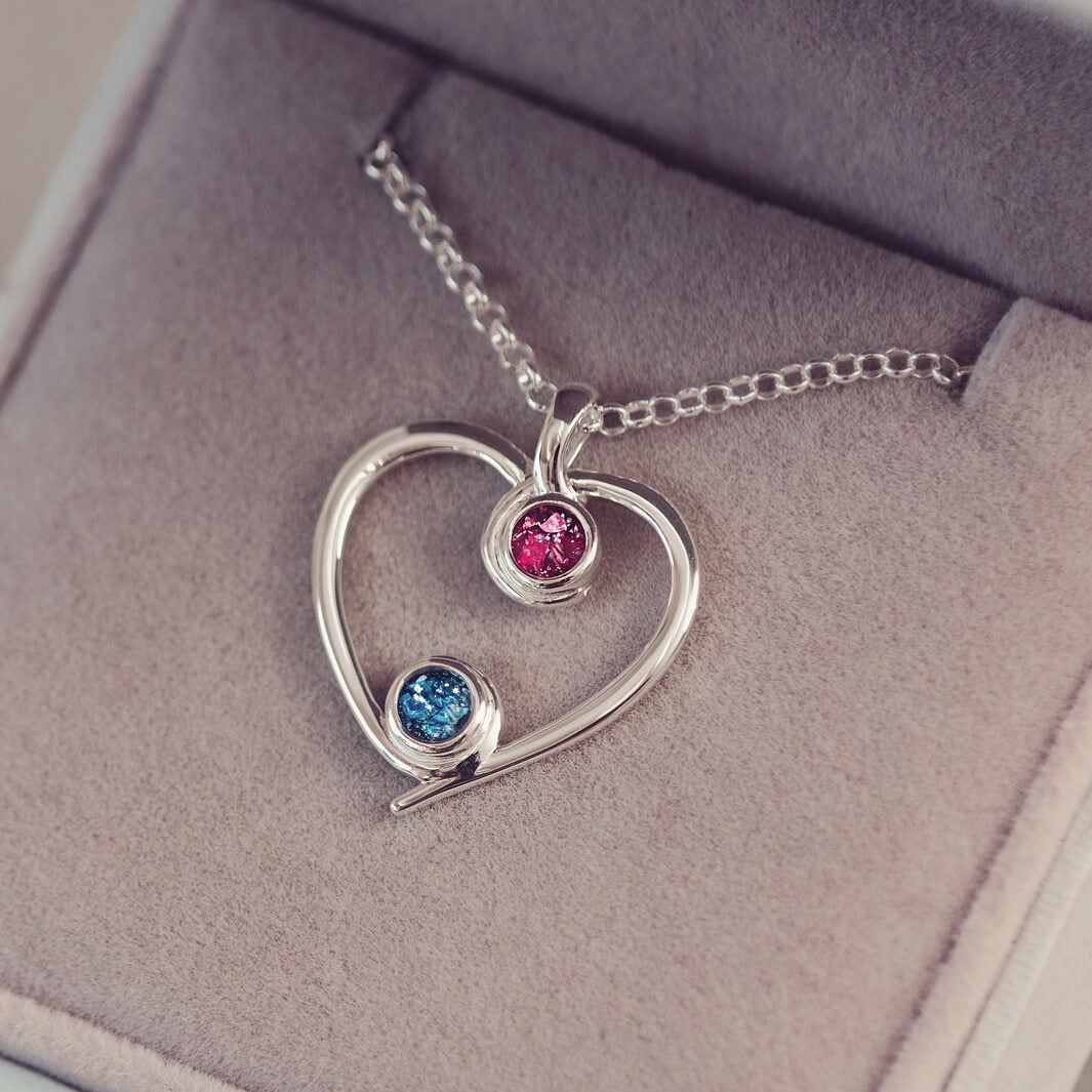 A silver heart-shaped pendant necklace featuring two sparkling pink and blue resin stones, holding cremation ashes, displayed against a neutral background.