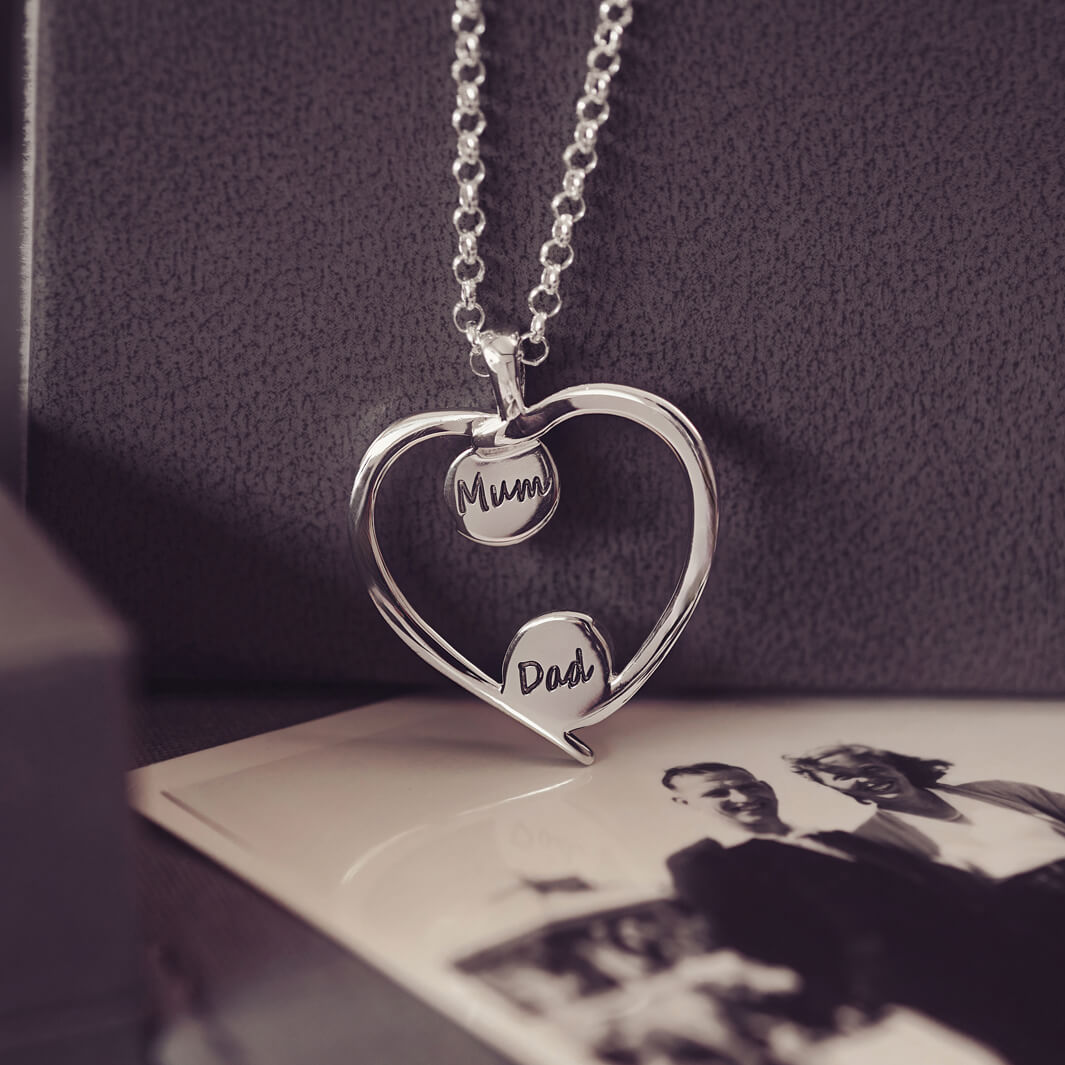 A silver heart-shaped pendant necklace featuring two short names (mum and dad) engraved onto the reverse of each bezel, holding cremation ashes, displayed against a neutral background.