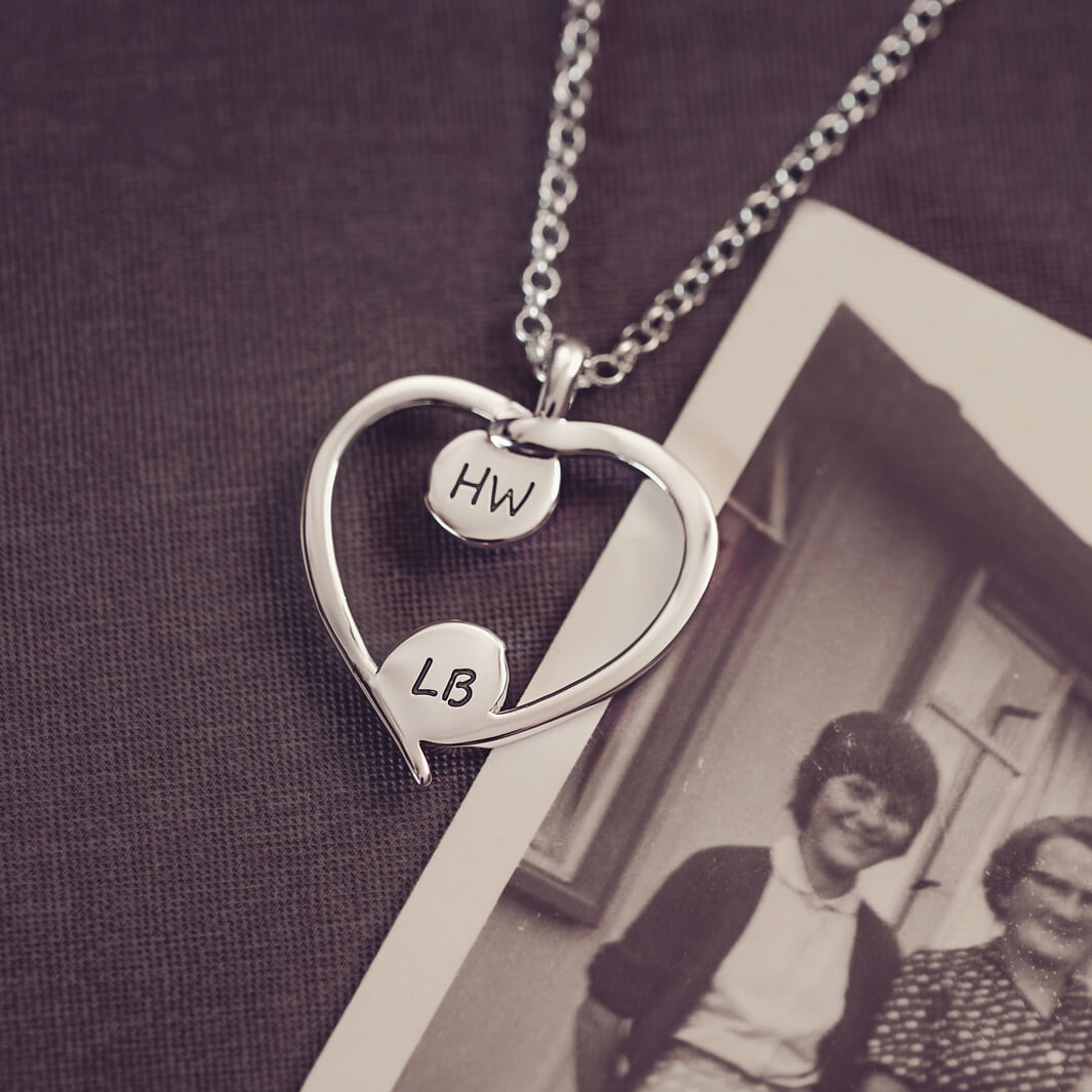 A silver heart-shaped pendant necklace featuring engraved initials on each bezel, holding cremation ashes, displayed against a neutral background.