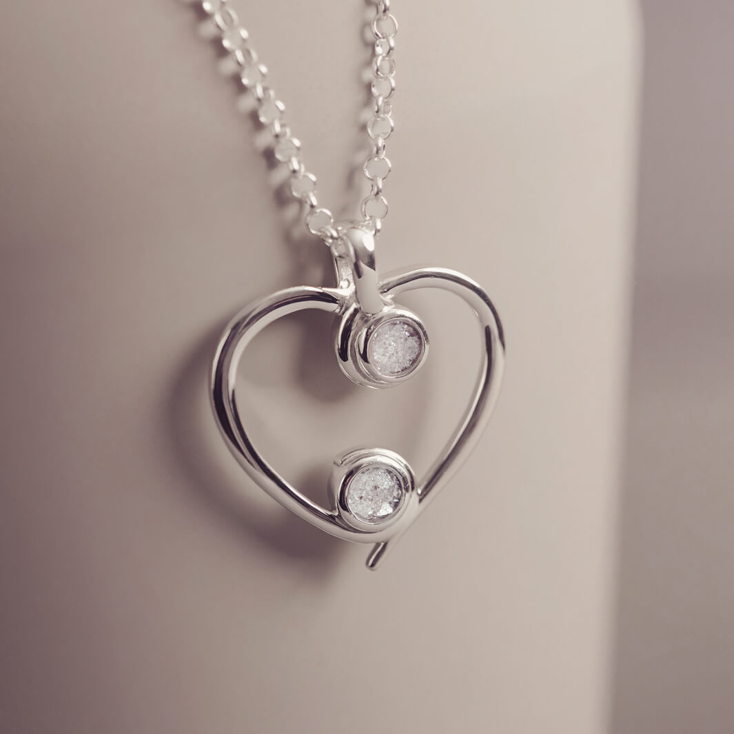 A silver heart-shaped pendant necklace featuring two sparkling clear resin stones, holding cremation ashes, displayed against a neutral background.