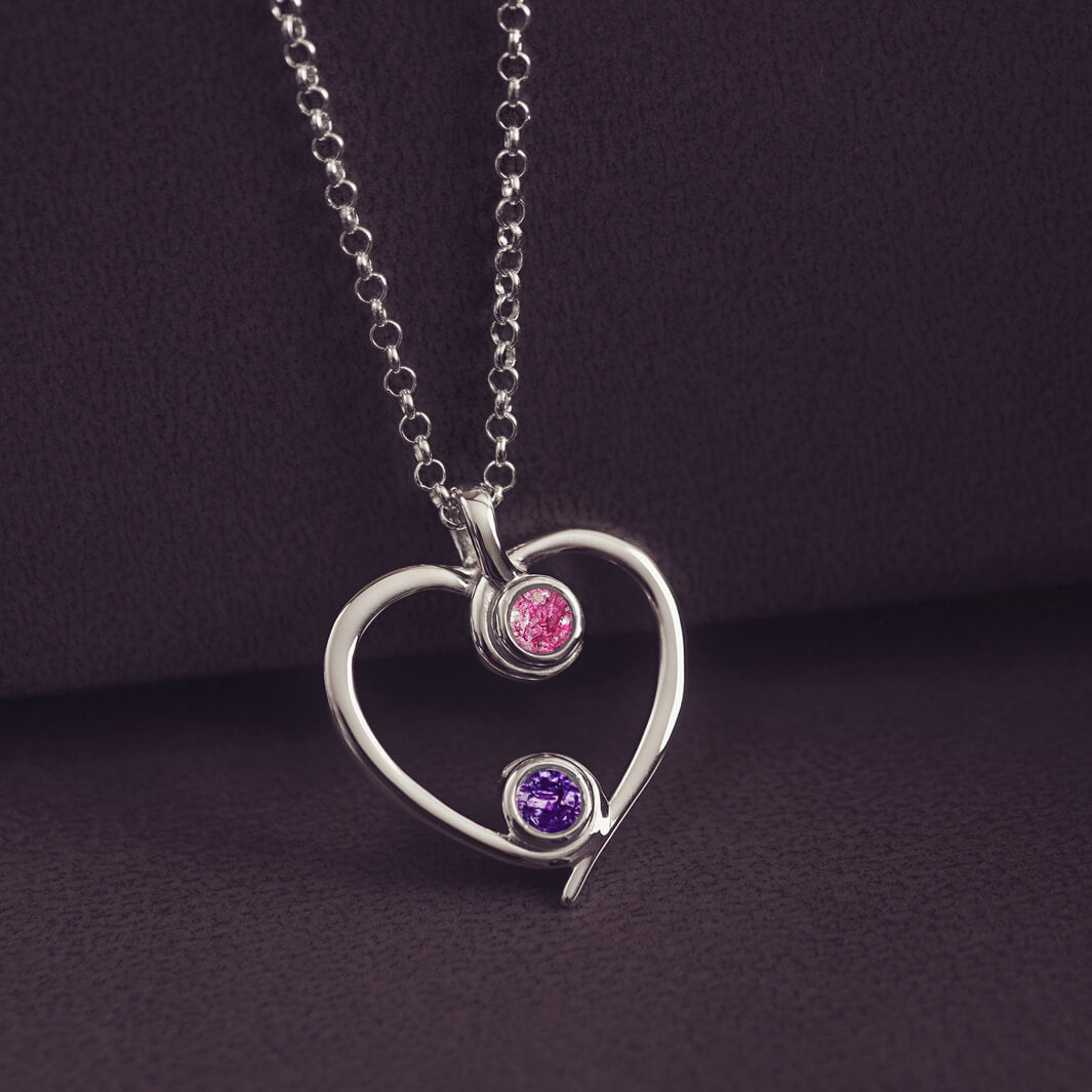 A silver heart-shaped pendant necklace featuring two sparkling pink and purple resin stones, holding cremation ashes, displayed against a dark background.