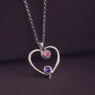 A silver heart-shaped pendant necklace featuring two sparkling pink and purple resin stones, holding cremation ashes, displayed against a dark background.