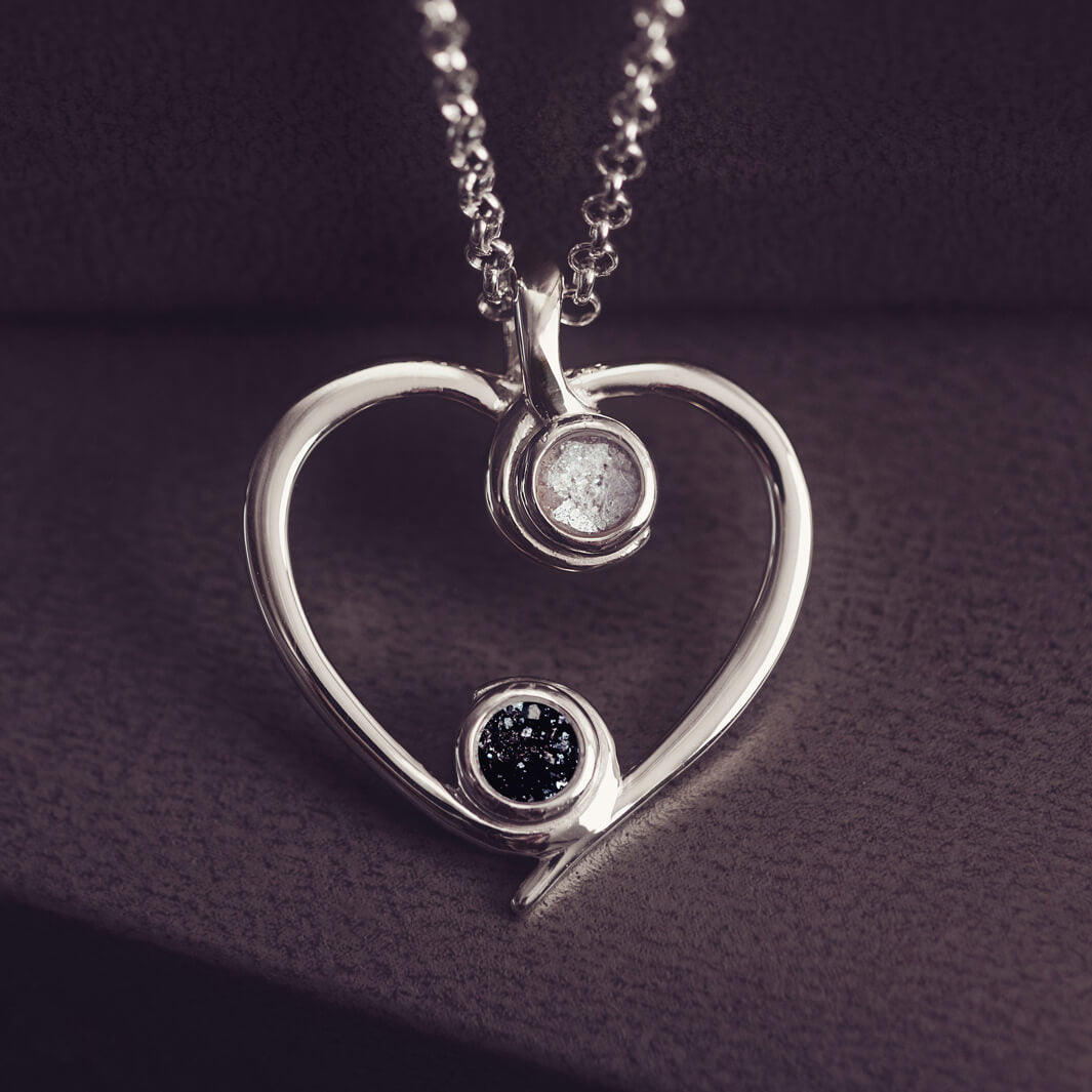 A silver heart-shaped pendant necklace featuring two sparkling white and black resin stones, holding cremation ashes, displayed against a dark background.