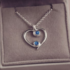 A silver heart-shaped pendant necklace featuring two sparkling blue resin stones, holding cremation ashes, displayed against a neutral background.