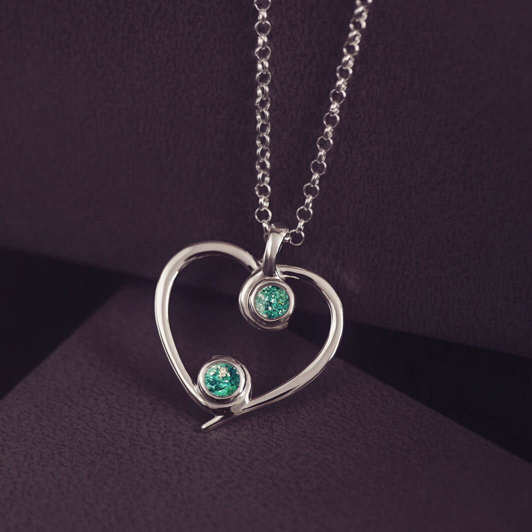 A silver heart-shaped pendant necklace featuring two sparkling green resin stones, holding cremation ashes, displayed against a dark background.