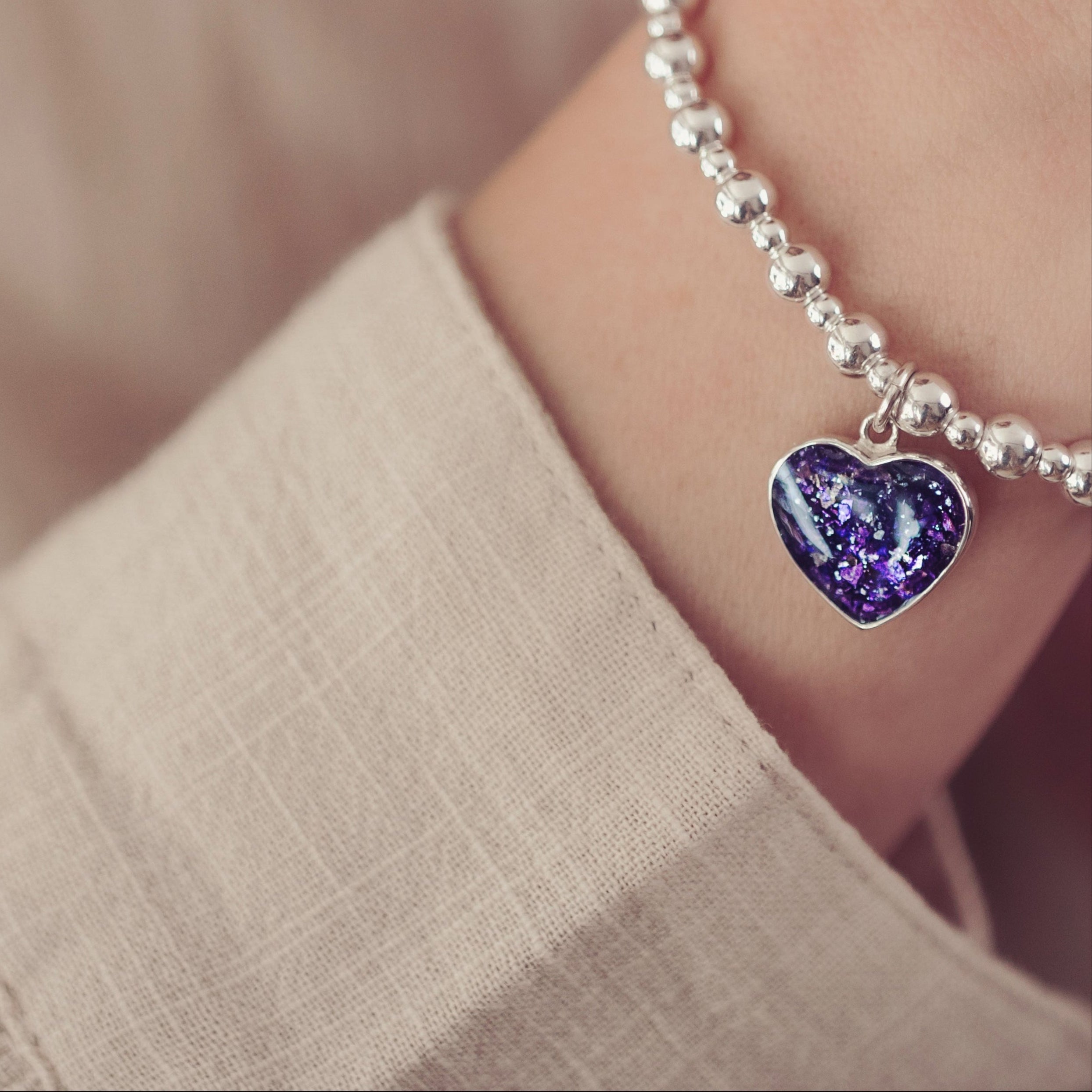 A close-up of a woman's wrist wearing a silver beaded bracelet with a purple heart charm containing cremation ashes