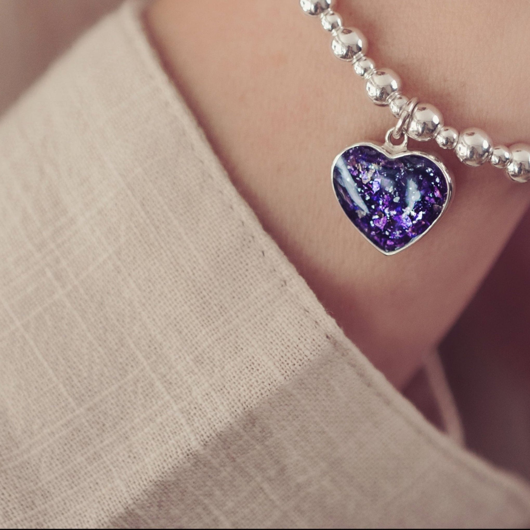 A close up of a woman's wrist wearing a silver beaded bracelet with a purple heart shaped charm, wearing a soft beige shirt in the background