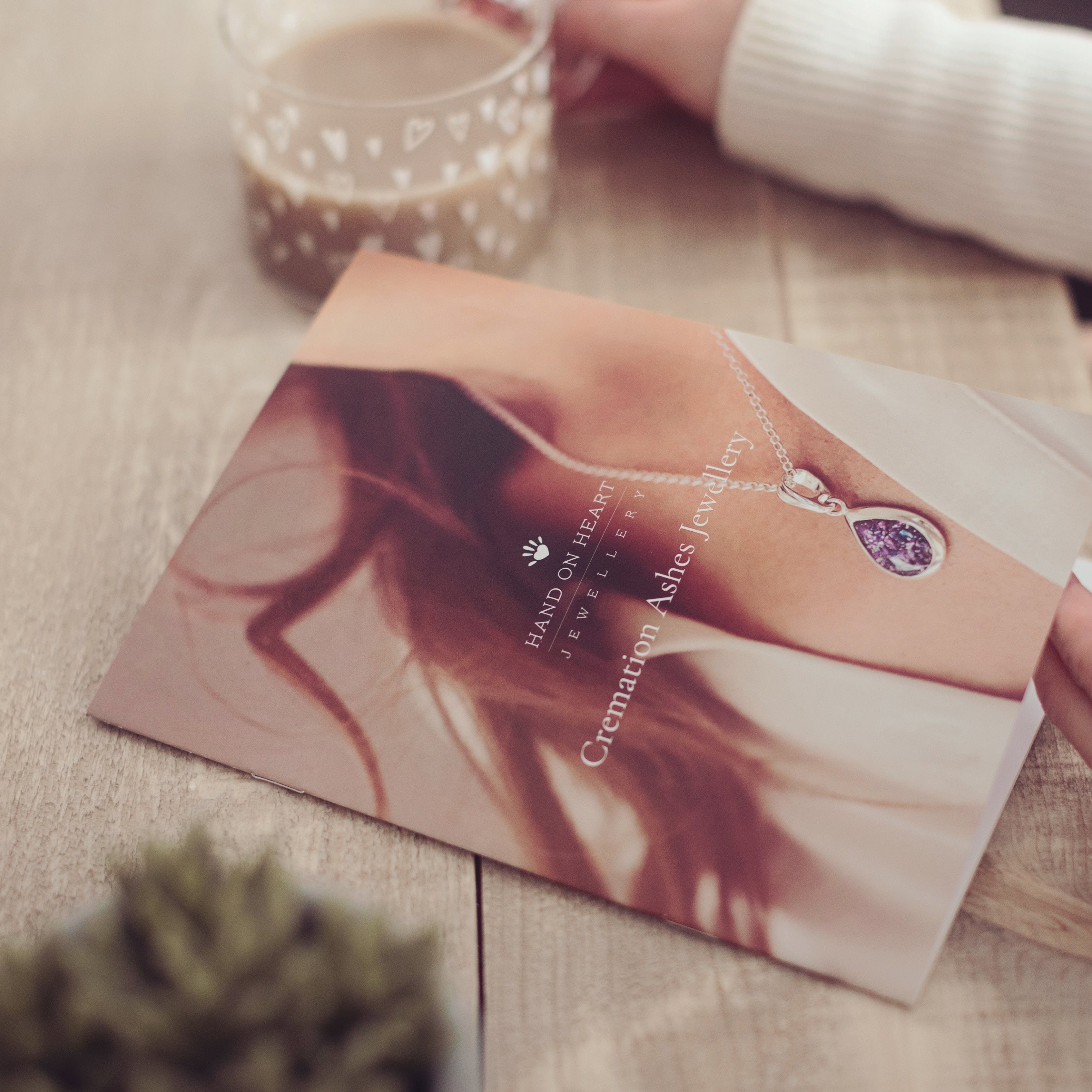 A woman holds our cremation ashes jewellery brochure with a glass cup of coffee blurred in the background