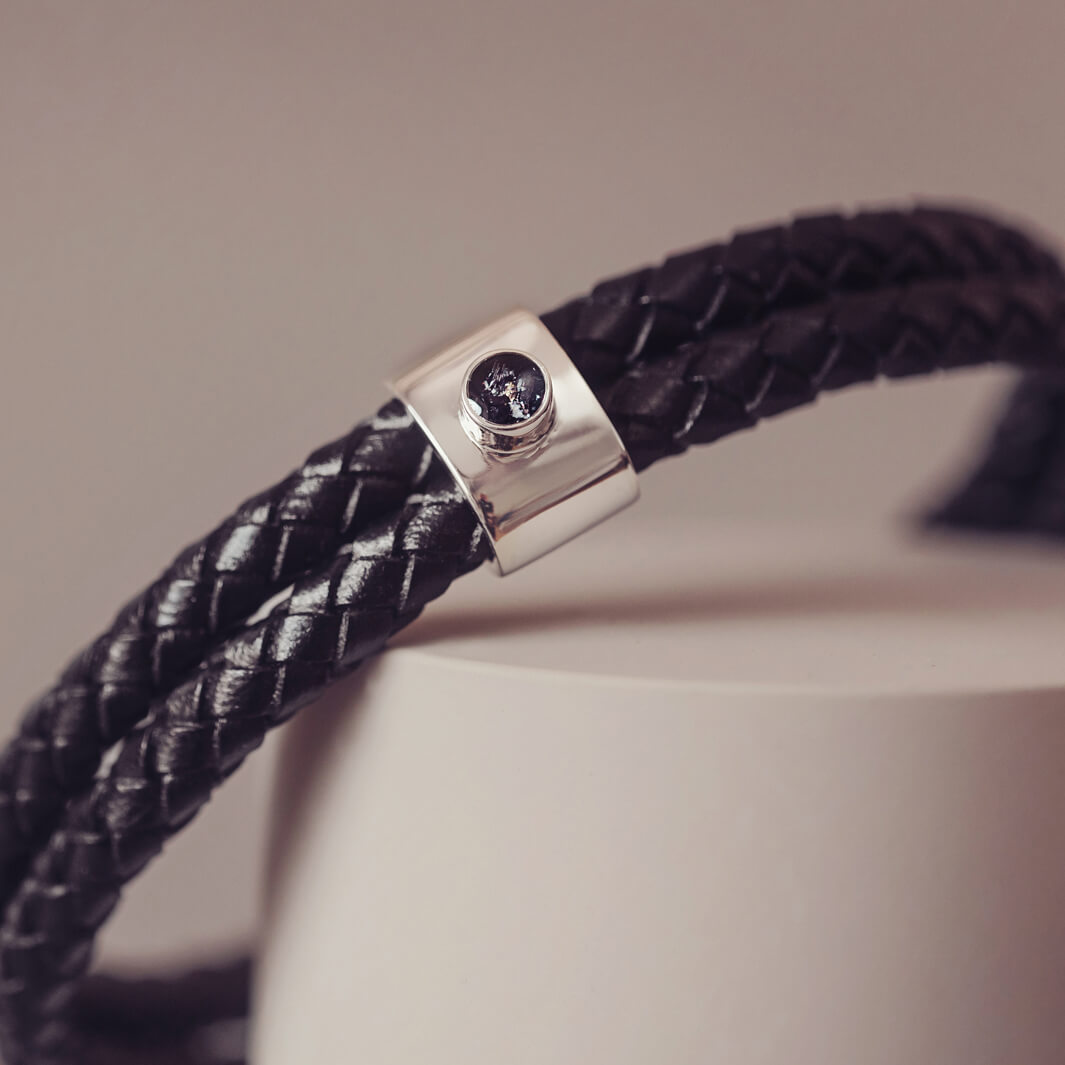 Black braided leather bracelet featuring a silver charm with a small black memorial stone set in the centre, displayed on a grey surface.