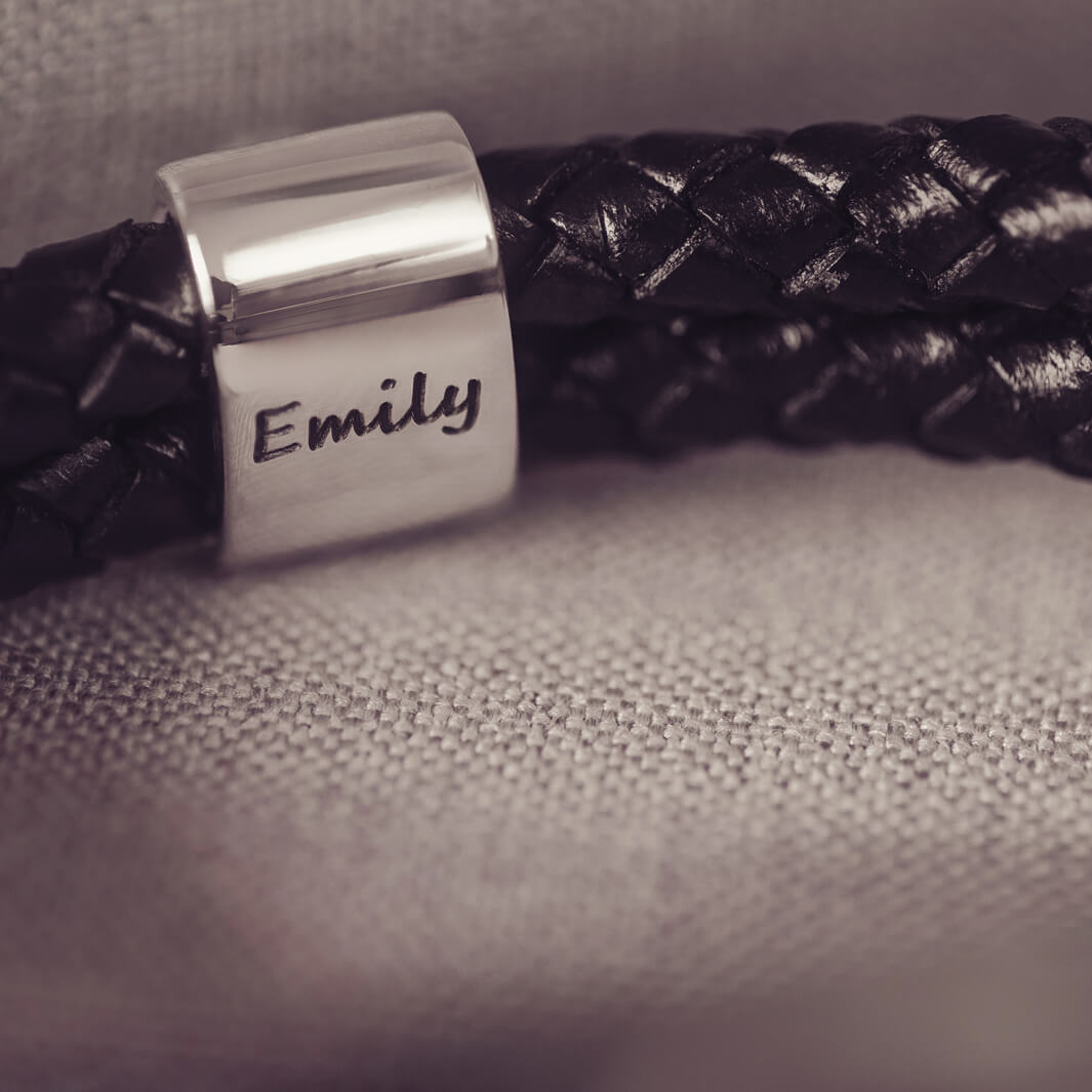 Black braided leather bracelet featuring a silver charm with the name 'Emily' engraved on it in black, displayed on a grey surface.