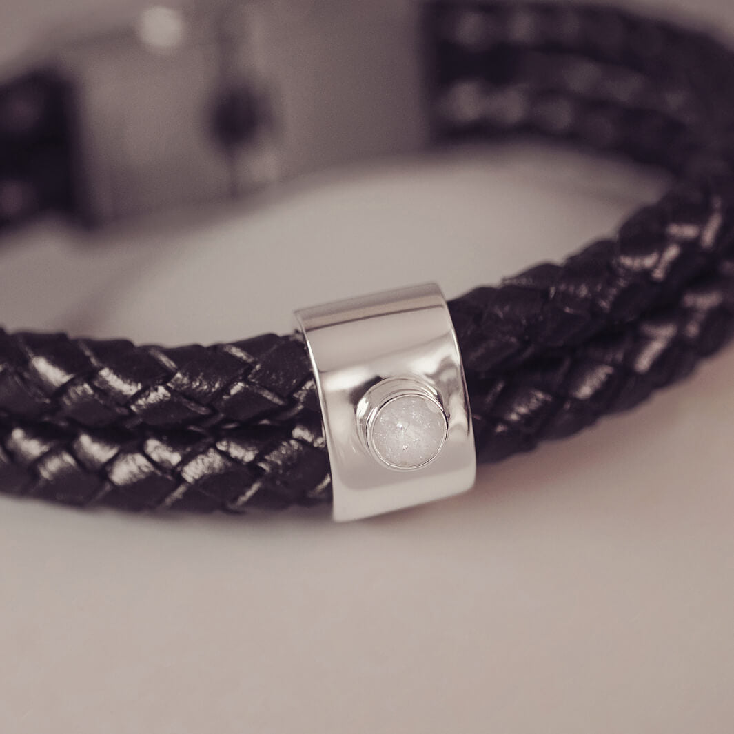 Black braided leather bracelet featuring a silver charm with a small clear memorial stone set in the centre, displayed on a grey surface.