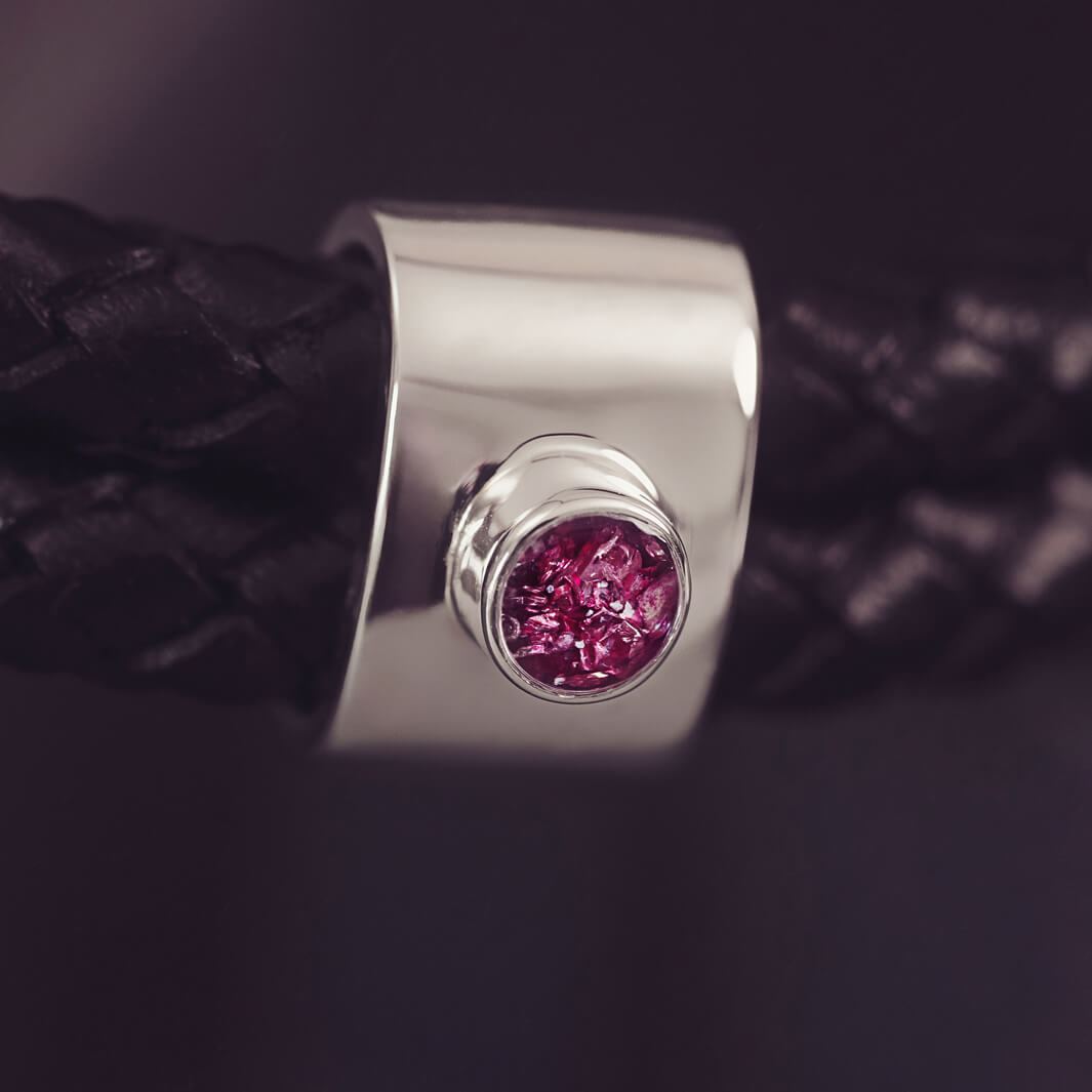 Black braided leather bracelet featuring a silver charm with a small pink memorial stone set in the centre, displayed on a grey surface.