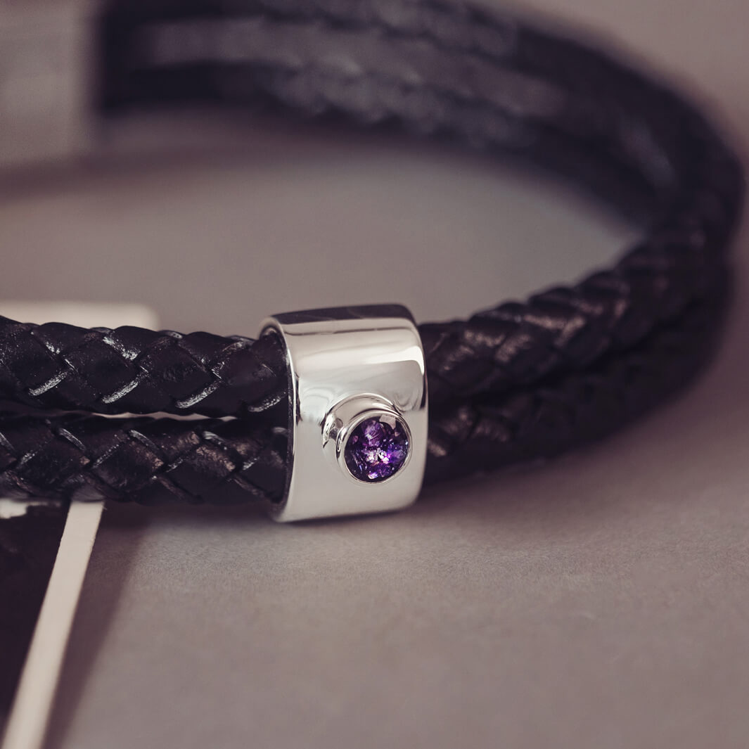 Black braided leather bracelet featuring a silver charm with a small purple memorial stone set in the centre, displayed on a grey surface.