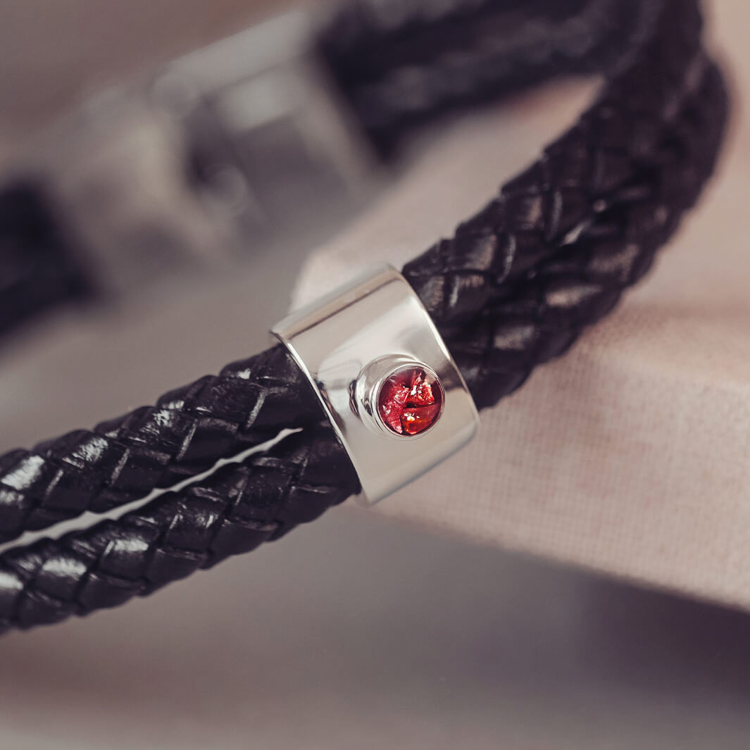 Black braided leather bracelet featuring a silver charm with a small red memorial stone set in the center, displayed on a grey surface.