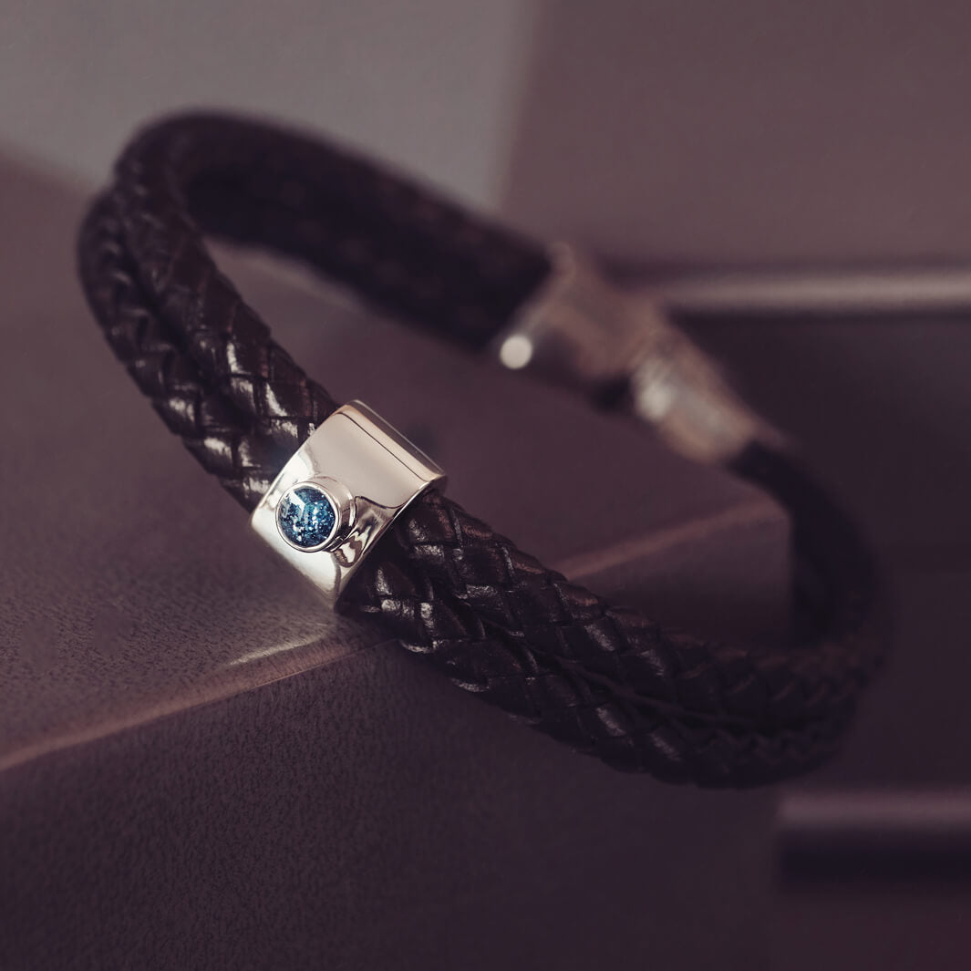 Black braided leather bracelet featuring a silver charm with a small blue memorial stone set in the centre, displayed on a grey surface.