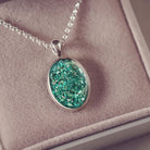 Sterling silver oval necklace with a central section filled with memorial ashes and green crushed crystals, displayed on a grey fabric background