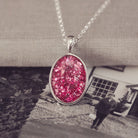 Sterling silver oval necklace with a central section filled with memorial ashes and pink crushed crystals, displayed on a background of a black and white photo, showing a lady stood in front of a house