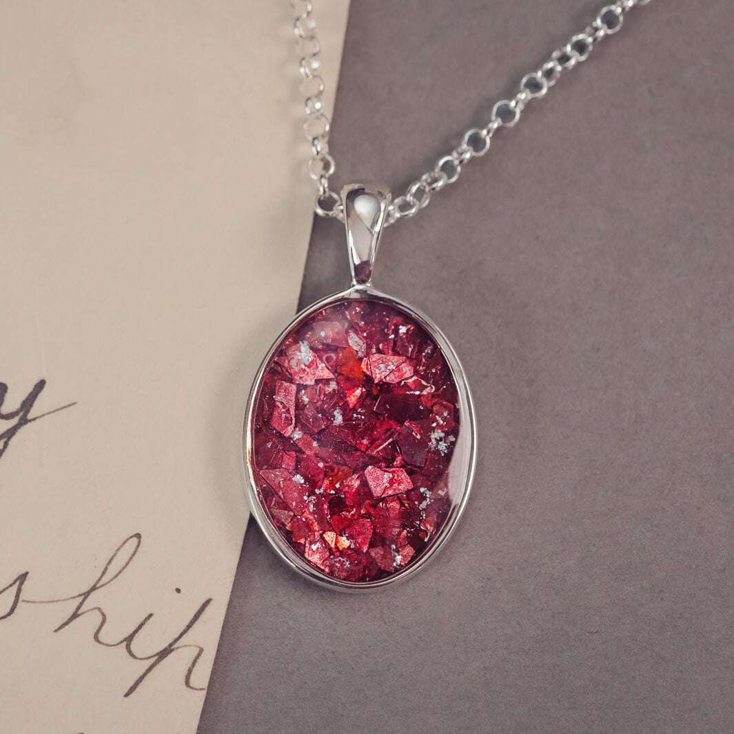 An oval shaped sterling silver pendant, on a silver belcher chain, filed with red crushed crystal and cremation ashes