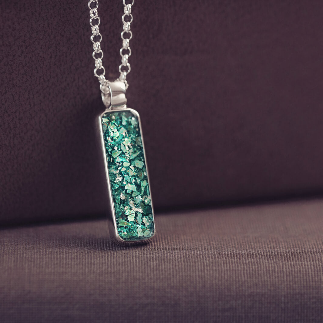 A sophisticated silver necklace featuring a rectangular pendant filled with shimmering green crystal and resin, delicately layered with cremation ashes, showcased against a dark background, on a silver belcher chain.