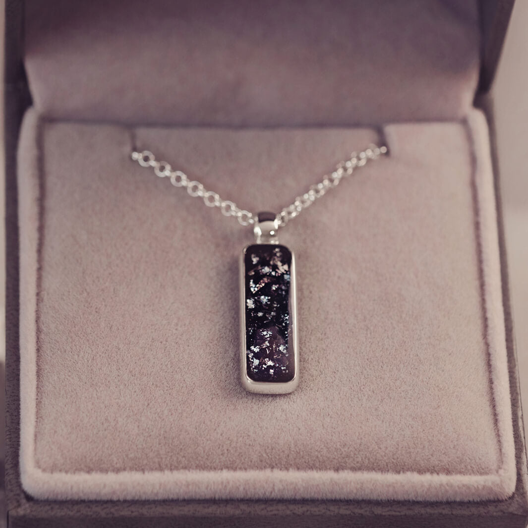 A sophisticated silver necklace featuring a rectangular pendant filled with shimmering black crystal and resin, delicately layered with cremation ashes, showcased against a light background, on a silver belcher chain.