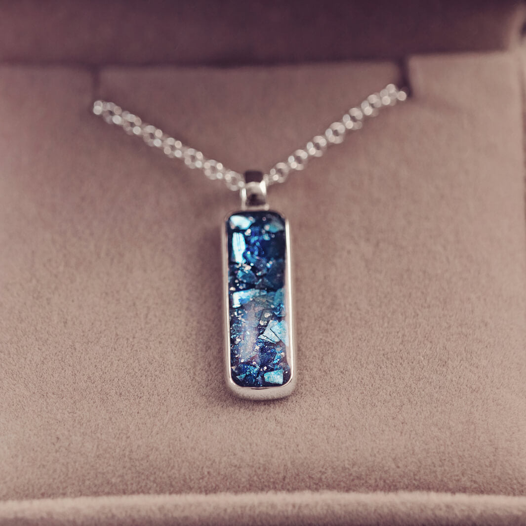 A sophisticated silver necklace featuring a rectangular pendant filled with shimmering blue crystal and resin, delicately layered with cremation ashes, showcased against a light background, on a silver belcher chain.