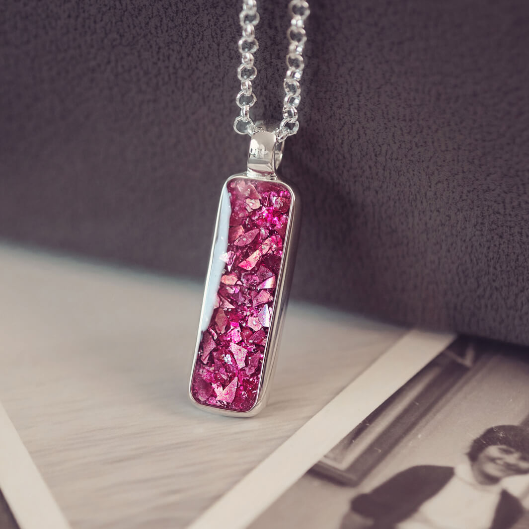 A sophisticated silver necklace featuring a rectangular pendant filled with shimmering pink crystal and resin, delicately layered with cremation ashes, showcased against a dark background, on a silver belcher chain.