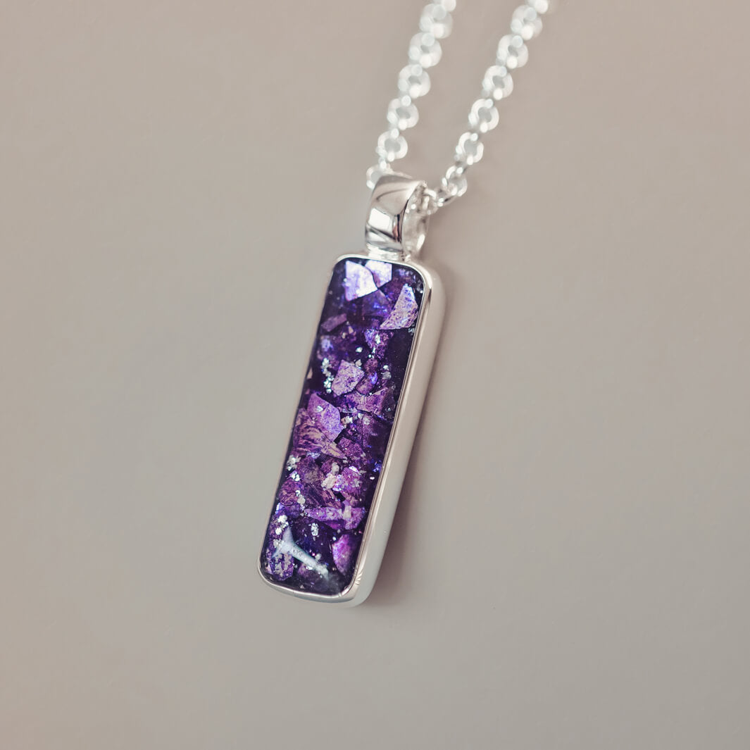 A sophisticated silver necklace featuring a rectangular pendant filled with shimmering purple crystal and resin, delicately layered with cremation ashes, showcased against a light background, on a silver belcher chain.