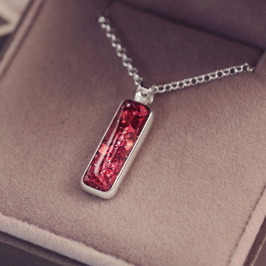 A sophisticated silver necklace featuring a rectangular pendant filled with shimmering red crystal and resin, delicately layered with cremation ashes, showcased against a dark background, on a silver belcher chain.