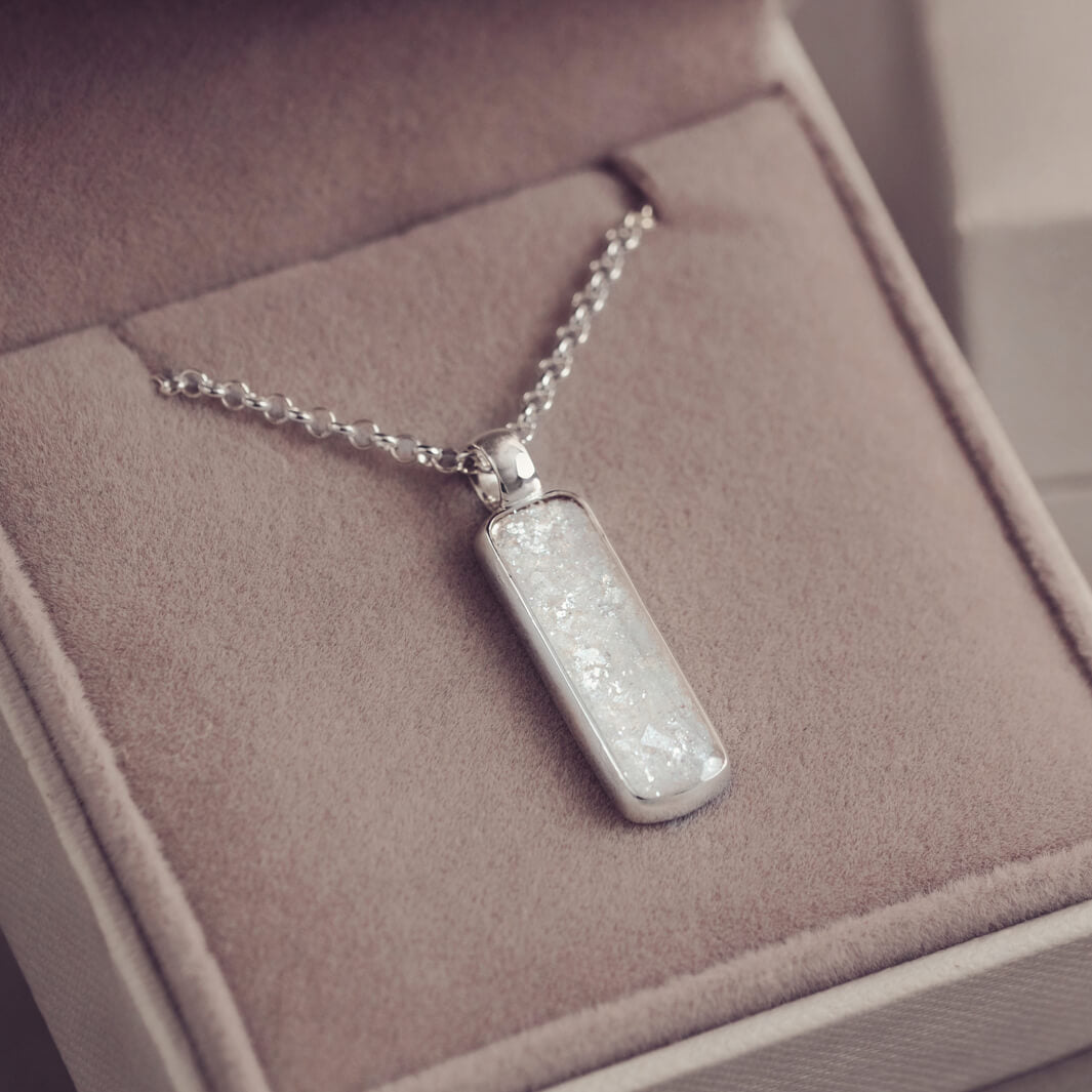 A sophisticated silver necklace featuring a rectangular pendant filled with shimmering white resin, delicately layered with cremation ashes, showcased against a light background.