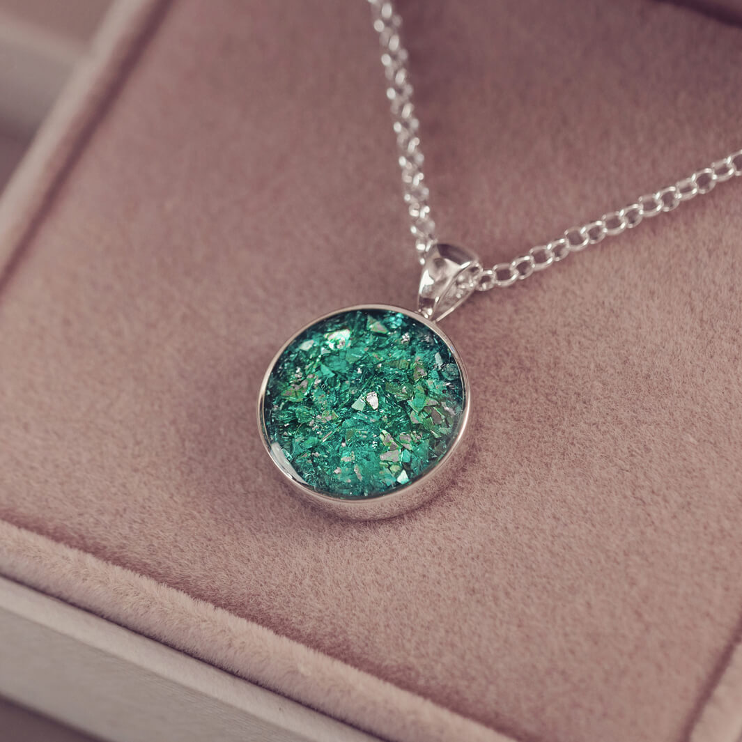 A round sterling silver necklace charm, hanging on a sterling silver chain. The charm is filled with green crystal and cremation ashes